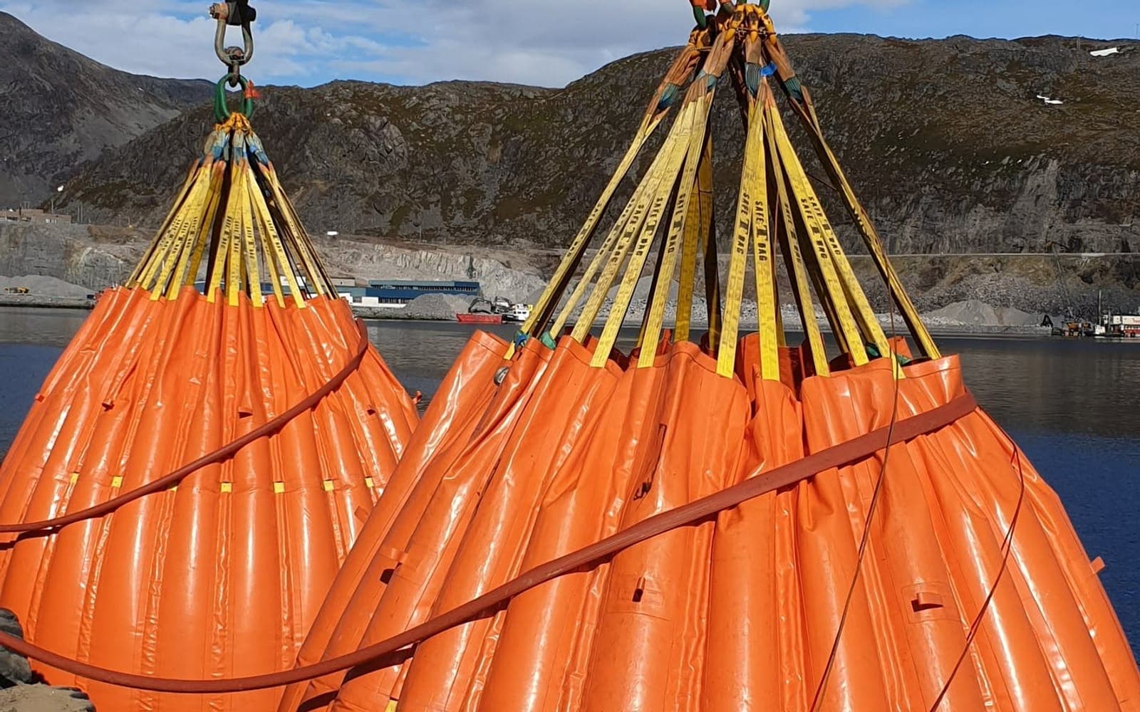 To oransje vannbagger henger i gule stropper på kaia under en løftetest, med fjell og fjord bak