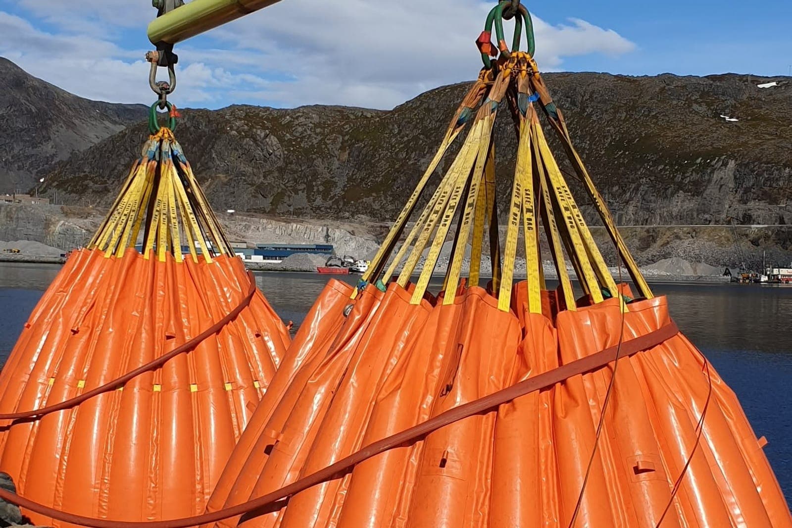 To oransje vannbagger fylt til maks henger i stropper under en gul løfteåk på kaia, med fjell og fjord bak
