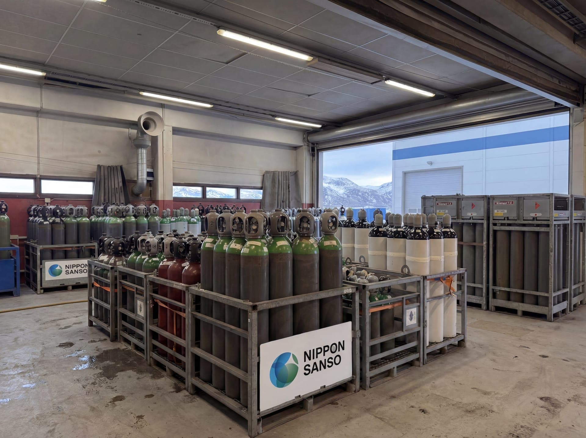 Rack med gassflasker inne i lagerhallen, med porten åpen mot fjellene